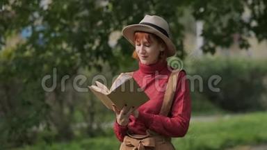 年轻迷人的红头发女人戴着帽子和毛衣在一个自然公园看书。 秋天凉爽的一天。